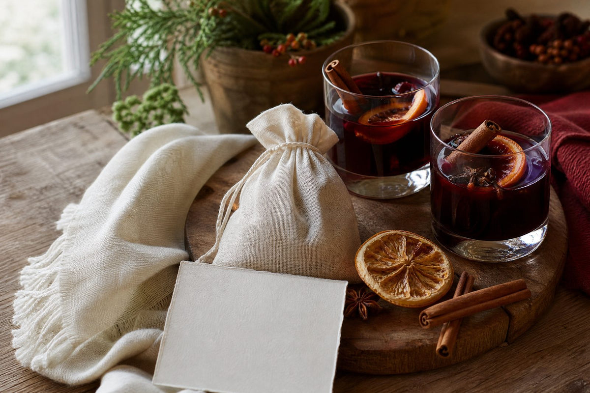 A completed, gift-ready mulled-drink hostess bundle: two warm glasses of mulled wine or spiced cider beside an unbranded muslin spice sachet, dried