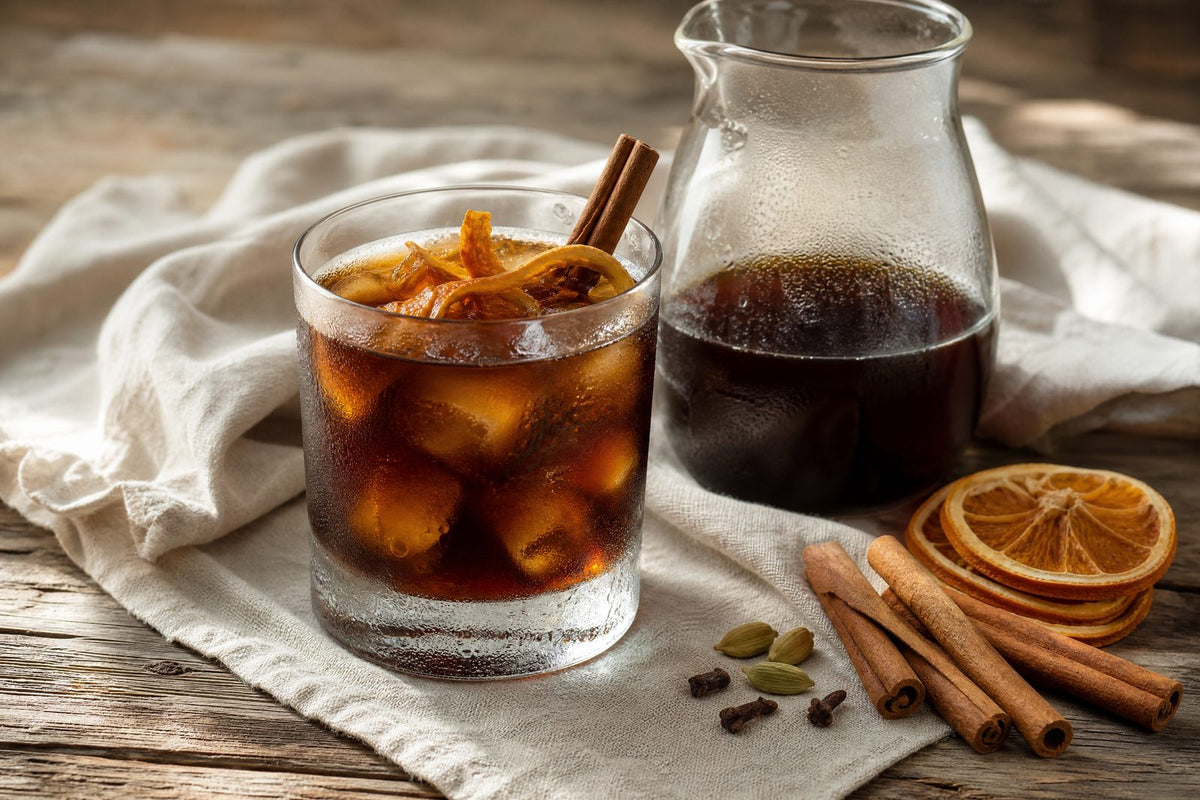 An ingredient-led editorial still life showing a finished cinnamon-orange spiced cold brew alongside the exact whole spices named in the recipe: cinnamon